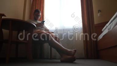 一个戴眼镜的男人坐在酒店房间窗户附近的椅子上，拿着电话。
